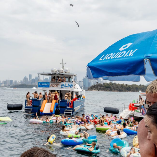 A large group of people enjoy a float party in a bay, with colorful inflatables in the water near boats. One boat displays "LIQUID I.V." branding. City skyline and birds are visible in the background.