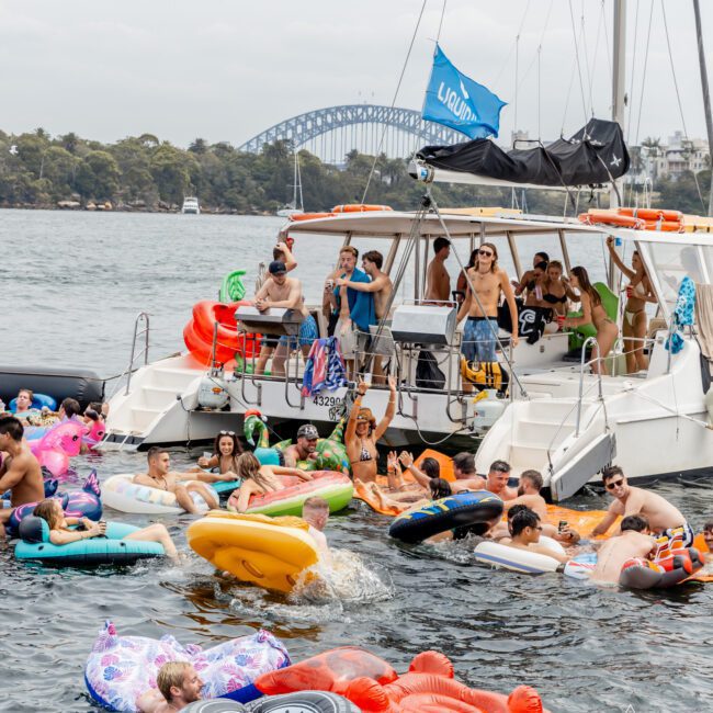 A group of people party on a boat and colorful inflatables in the water, with the Sydney Harbour Bridge visible in the background on a cloudy day. The scene is lively and festive.