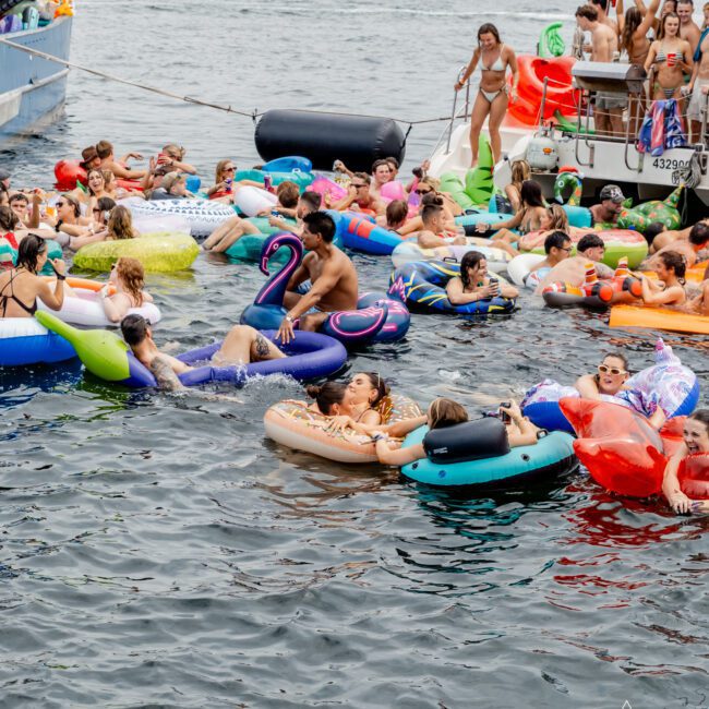 A large group of people on colorful inflatable floaties relax and socialize in the water near boats, with a forested shoreline and city skyline visible in the distance. The atmosphere appears festive and lively.
