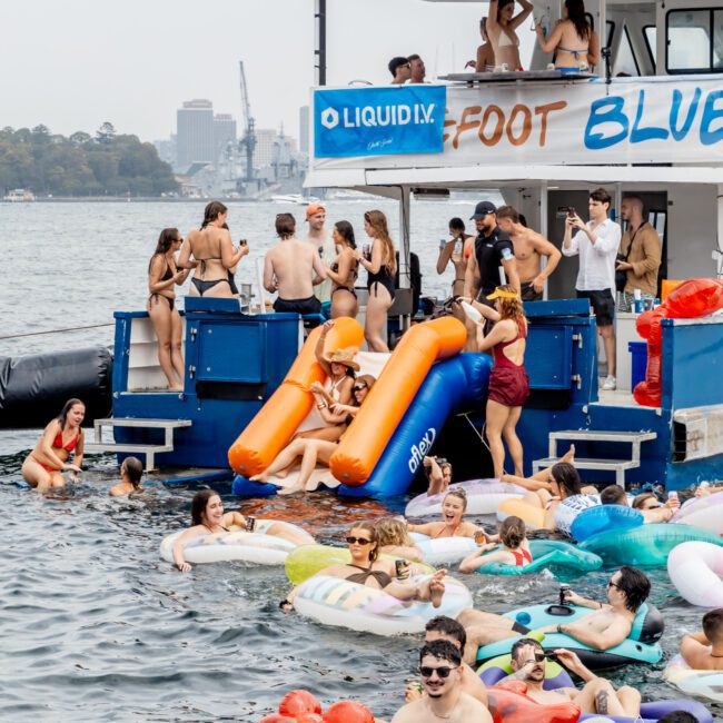 A lively group of young people on a boat party and in the water on floats, with some using an orange slide from the boat into the river. The atmosphere is festive and summery.