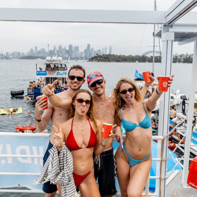 Four young adults in swimwear, smiling and holding red cups, pose on a dock with boats and a city skyline in the background. It's a lively summer scene by the water, suggesting a festive or party atmosphere.