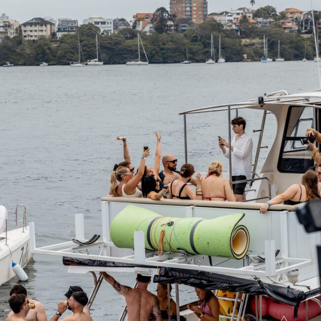 A group of people in swimsuits are partying on a docked boat, some raising their arms and posing for a photo, with water, sailboats, and city buildings visible in the background.