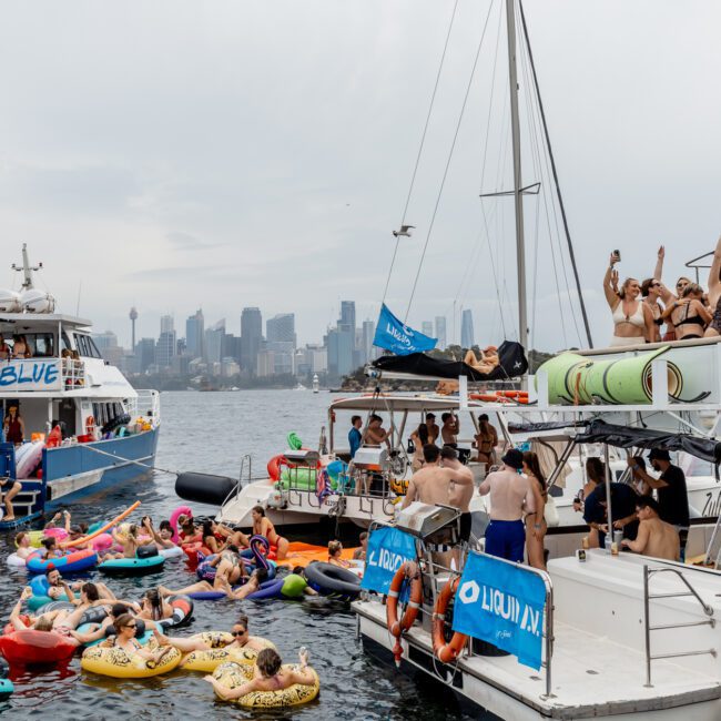 Dozens of people in swimsuits enjoy a party on boats and colorful inflatables in the water, with a city skyline visible in the background under a cloudy sky.