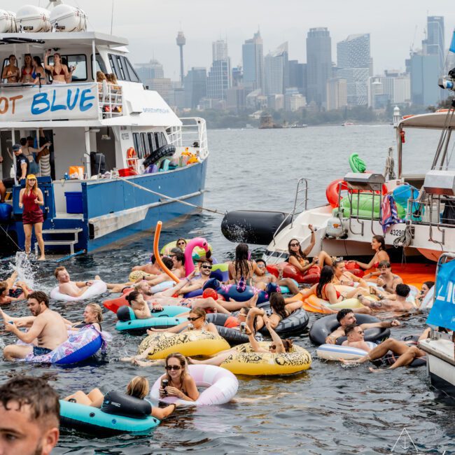 A lively group of people relax on colorful inflatable floats in the water near two docked boats, with a city skyline and cloudy sky in the background. Some people are on the boats, and others swim or float nearby.