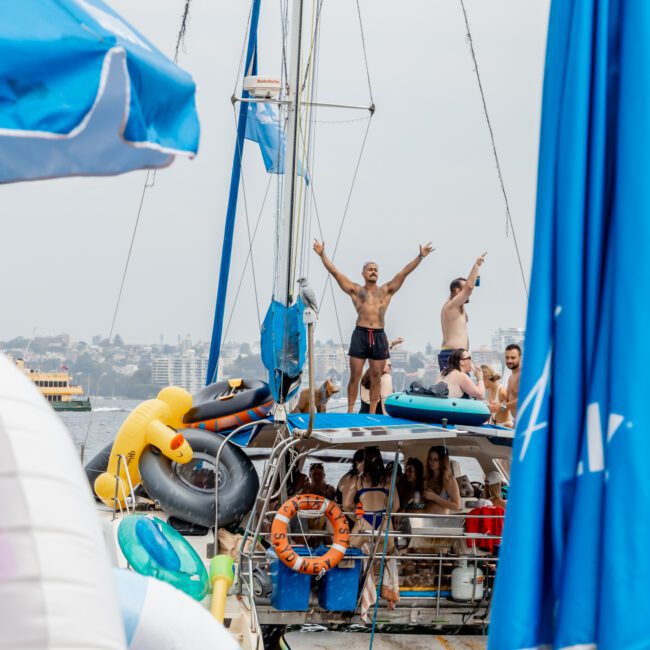 A group of people on a boat party; some stand on the deck, two men pose with arms raised on the roof. Inflatable floaties and blue umbrellas surround the boat, with a city skyline visible in the background.