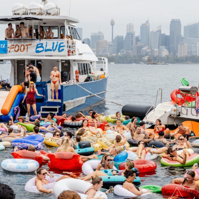 A large group of people float on colorful inflatables in the water near boats, with a city skyline visible in the background under a cloudy sky.