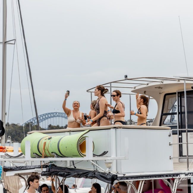 A group of women in swimsuits stand on the upper deck of a boat, taking selfies and holding drinks. Rolled-up mats are attached to the railing, and the Sydney Harbour Bridge is visible in the background.