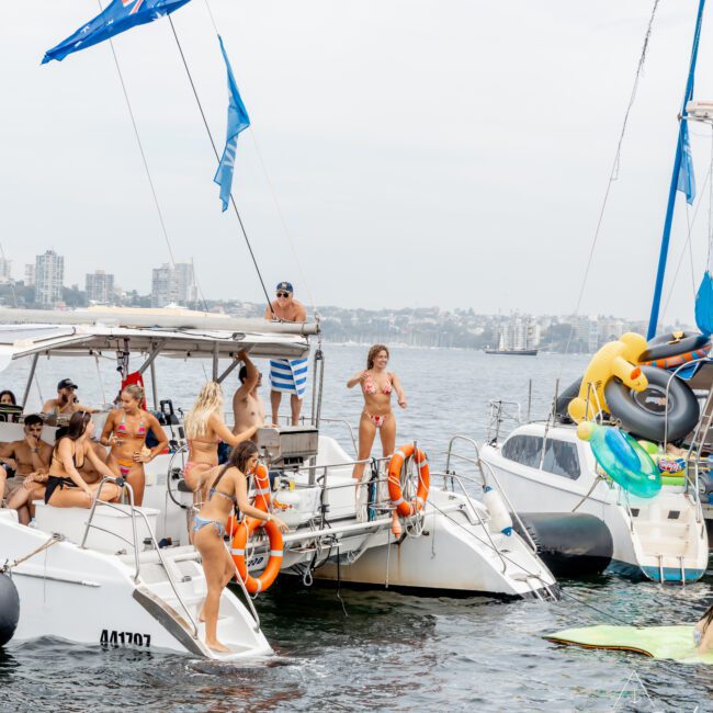 A group of people in swimsuits gather on two anchored boats on the water, with city buildings in the background. Some are standing and chatting, and inflatable toys and blue flags are visible. The scene appears lively and social.
