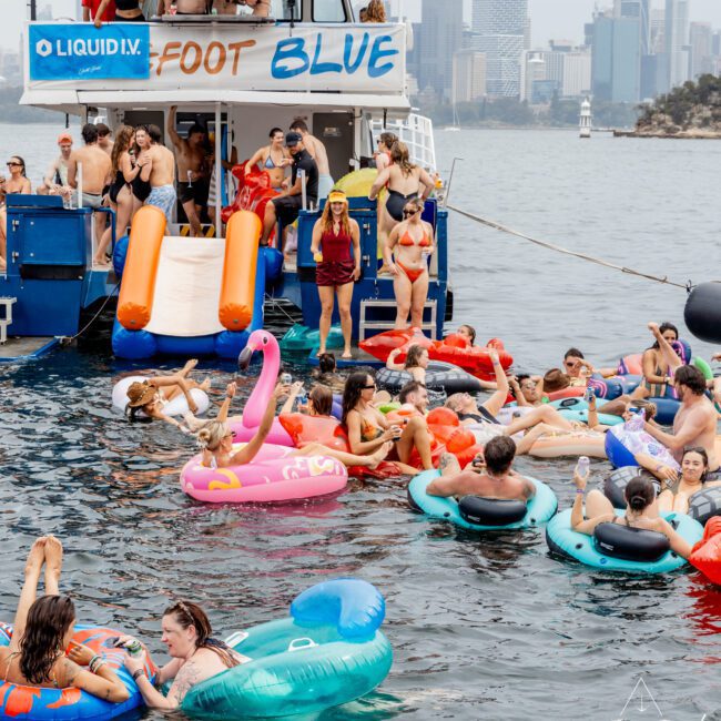 A lively group of people enjoy a yacht party, relaxing on colorful pool floats in the water. A large yacht named "Foot Blue" is anchored nearby, with people socializing on board. The city skyline is visible in the background.