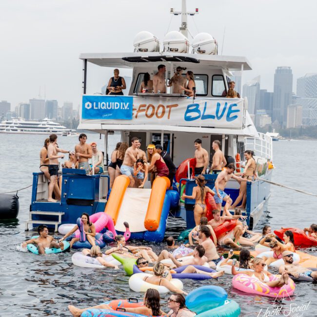 A lively group of people in swimsuits enjoy a party on a boat and in the water with colorful floaties. The boat has a “FOOT BLUE” sign, a slide, and city skyscrapers in the background.