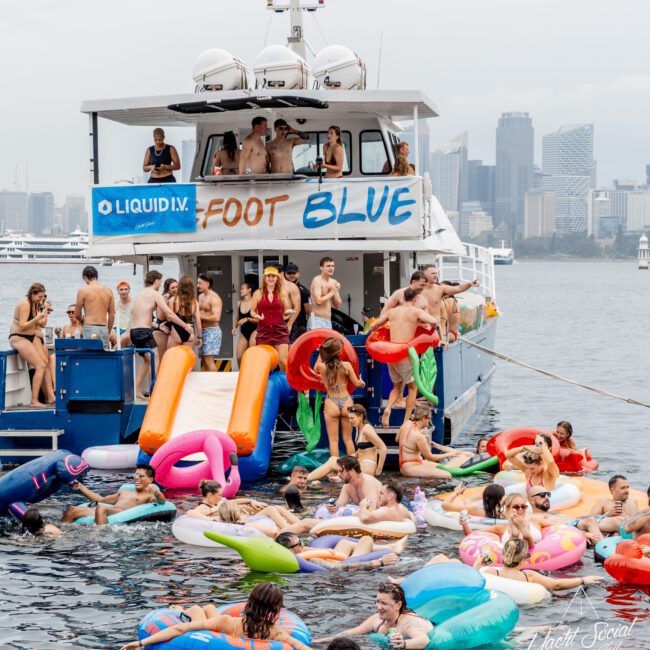 A group of people in swimsuits enjoy a lively pool party on and around a boat with inflatable toys. Some are on the boat, others float in the water on colorful inflatables, with a city skyline in the background.