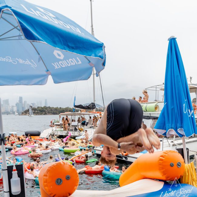 A person in swimwear does a backflip off an inflatable orange and blue platform into the water, with a crowd of people relaxing on colorful floaties nearby, and city buildings visible in the background.