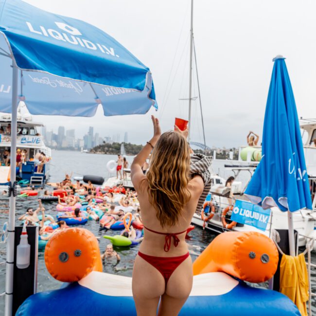 A woman in a red bikini stands on a float, raising a cup toward a lively crowd on inflatables and boats at a waterfront party, with city buildings and overcast sky in the background.