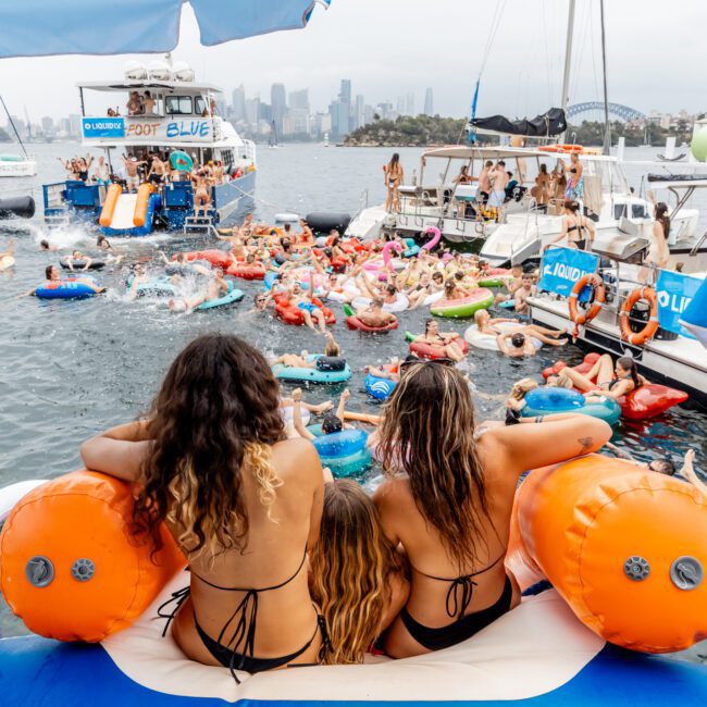 Three women sit on an inflatable float at the edge of a boat, overlooking a lively party with people swimming, lounging on floats, and gathering around boats in a harbor, with a city skyline in the background.
