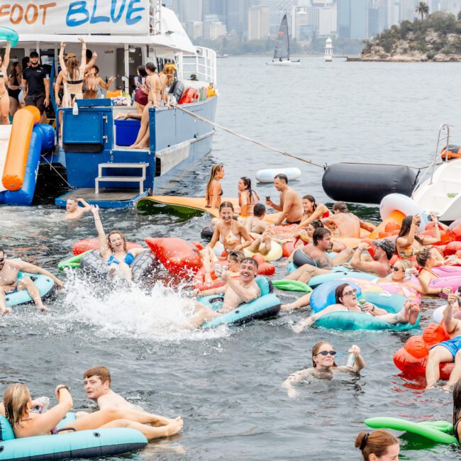 A lively group of people float on colorful pool inflatables in the water near a boat named "FOOT BLUE," with a city skyline visible in the background under a cloudy sky.