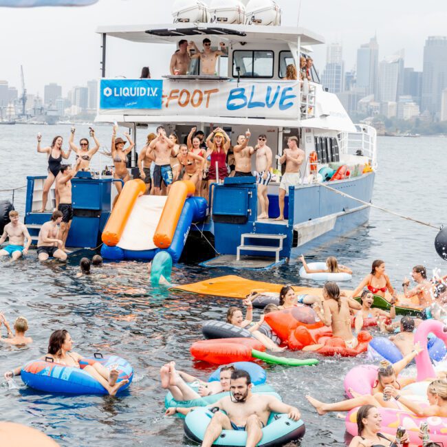 A lively party scene on the water with people on a boat and many others in colorful inflatables in the water. The boat, named "Foot Blue," has slides, and people are dancing and having fun under a cloudy sky near a city skyline.
