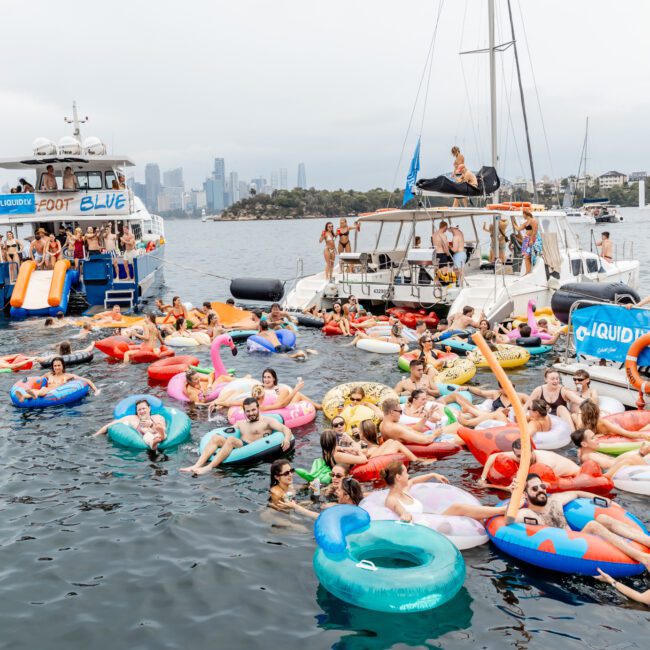 A lively scene of people on colorful inflatable pool floats in the water, surrounded by boats. The crowd is enjoying a party, with the city skyline and a cloudy sky visible in the background.