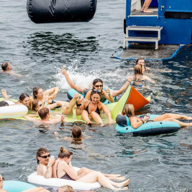A group of people relax on colorful inflatables in the water near a boat, laughing and enjoying themselves on a sunny day. Some are in swimsuits, others are splashing, and the atmosphere is lively and fun.