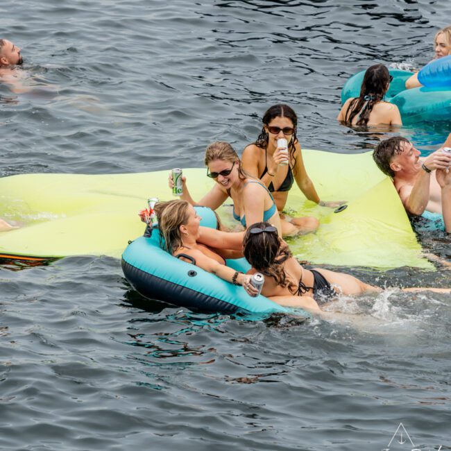 A group of people in swimsuits relax on colorful inflatables and swim in a body of water, socializing and holding drinks. The atmosphere is lively with several people enjoying the water together.