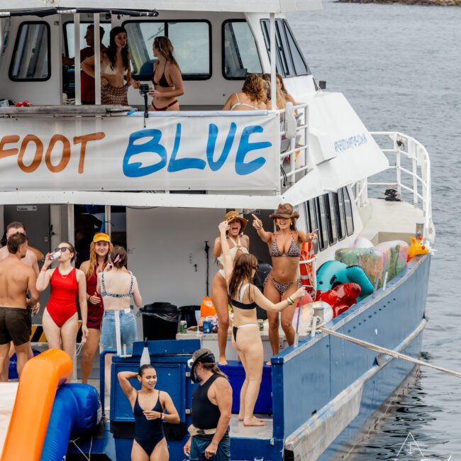 A group of people in swimwear enjoy a party on a large blue and white boat, with some standing, talking, and taking photos on deck. The boat is docked on a calm body of water near scenic buildings.