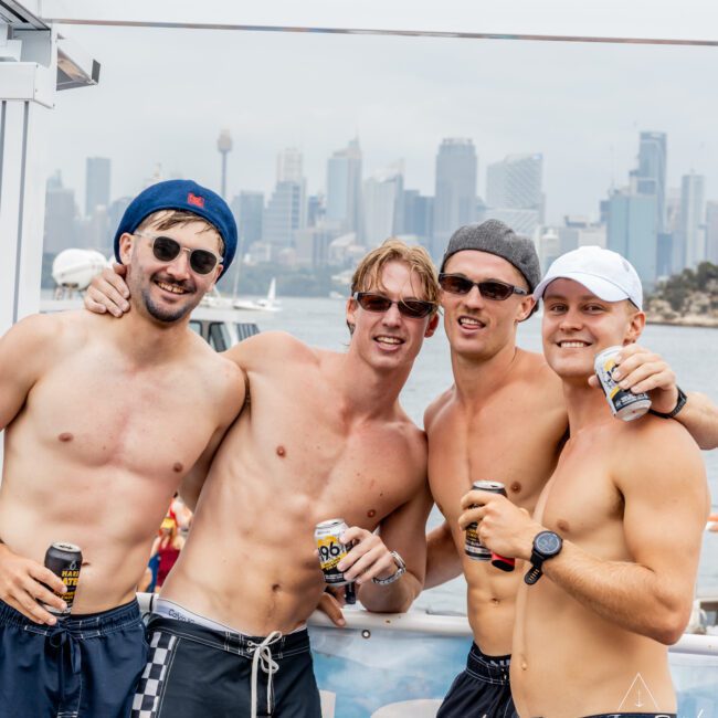 Four smiling, shirtless men in sunglasses and swim trunks stand arm-in-arm on a boat, holding drinks. The city skyline and water are visible in the background. The image has a “Yacht Social Club” logo in the corner.