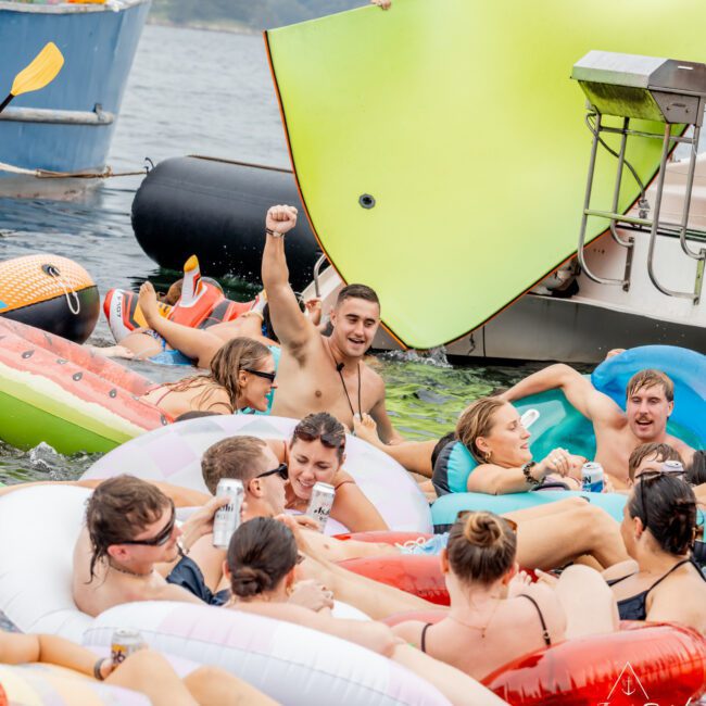 A group of people relax on inflatable pool floats in the water, smiling and holding drinks. A man in the center raises his fist in the air. Boats, large floats, and a city skyline are visible in the background.