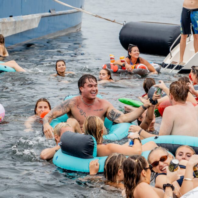 A group of people in swim rings enjoy a lively party in the water near a boat, smiling, talking, and raising drinks, surrounded by others swimming and floating on a sunny day.