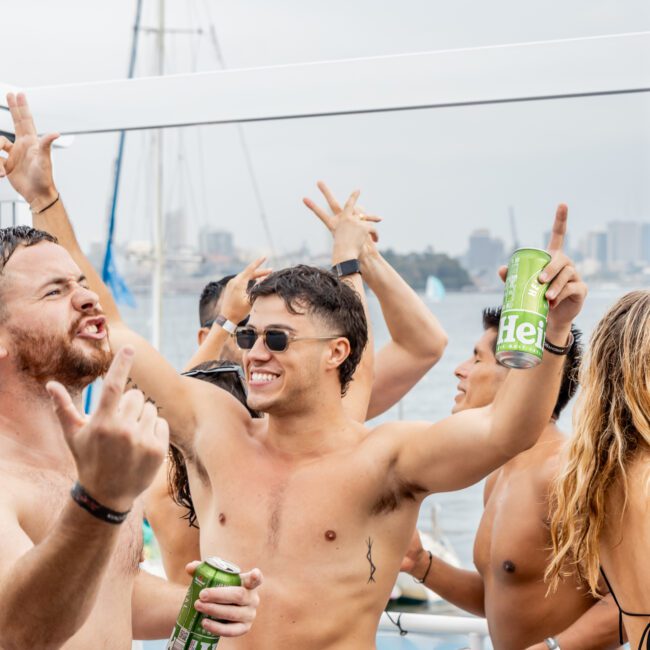 Shirtless people on a boat party cheer and hold drinks, with one person raising a fist. The group appears happy and energetic, with water, city buildings, and a flag visible in the background.