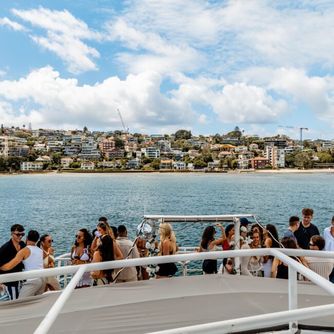 A group of people socializing on the deck of a yacht, with a coastal town and buildings visible in the background across the water under a partly cloudy sky.