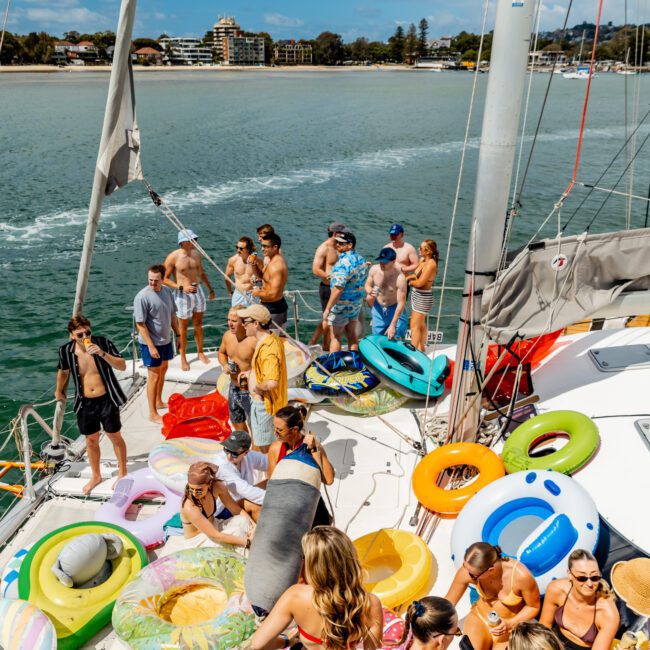 A group of people enjoy a sunny day on a boat, surrounded by colorful inflatable pool floats, chatting and relaxing with water and drinks, with a scenic shoreline in the background.