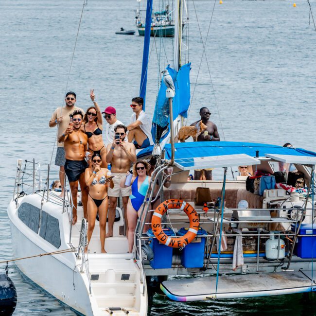 A group of people in swimsuits smile and pose on a sailboat in the water, some holding drinks. The boat has a blue canopy, and buildings and a bridge are visible in the background on a sunny day.