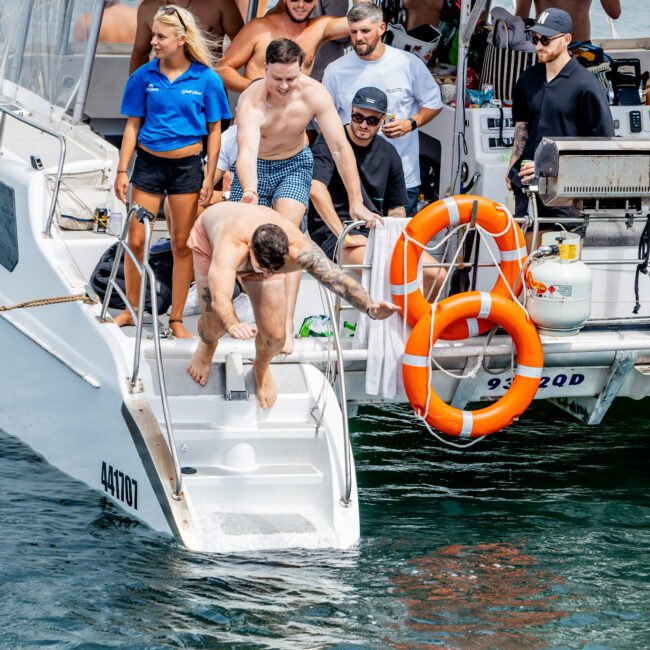 A group of people on a boat are smiling and watching as a man in swim trunks dives into the water from the boat’s rear steps. Bright orange life rings and boat equipment are visible.