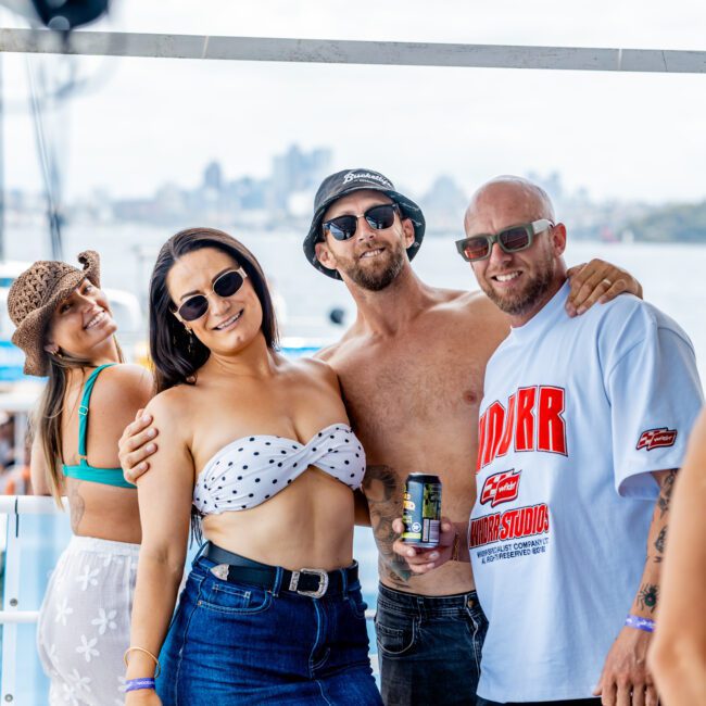 Four adults in casual summer outfits pose and smile on a boat, with water and a city skyline in the blurry background. One man holds a drink can. The atmosphere is cheerful and relaxed.