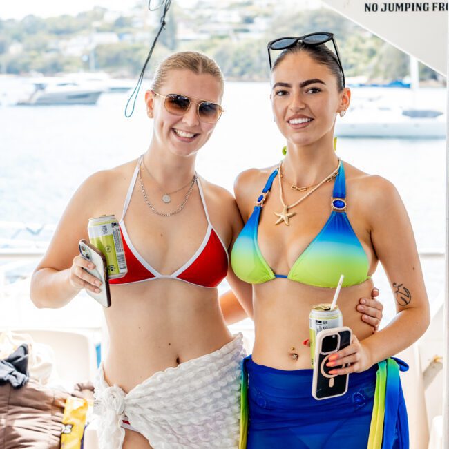 Two women in colorful bikinis and sunglasses smile while holding drinks on a boat, with water and boats visible in the background on a sunny day.