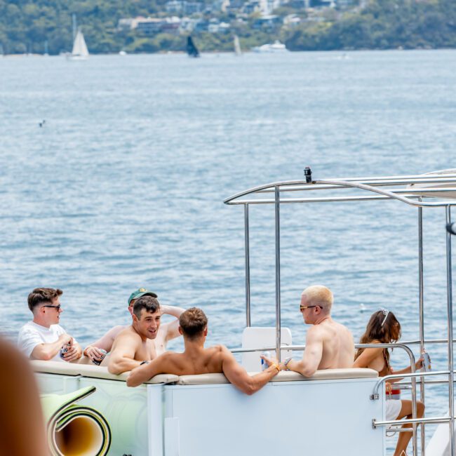 Five people relax on a boat on the water, talking and enjoying the sunny weather. A shoreline with houses and trees is visible in the background, along with sailboats on the water.