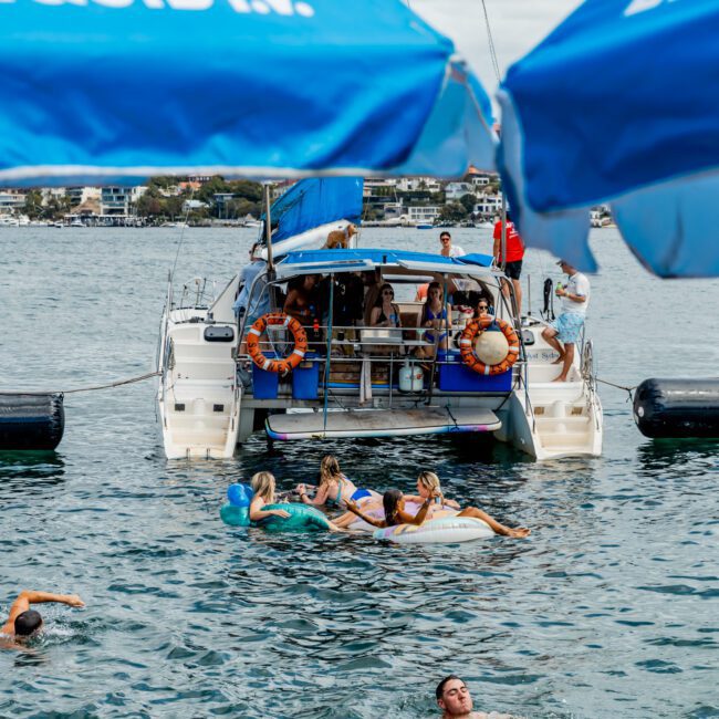 People relax on a catamaran and swim in the ocean, with several floating near the boat. Blue umbrellas with “LIQUID I.V.” branding are visible at the top of the image. A coastline is visible in the background.