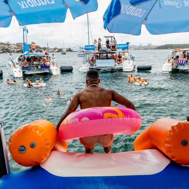 A person with a pink and orange inflatable ring slides into the water from a yacht, surrounded by floating people and anchored boats at a lively outdoor party under blue umbrellas.