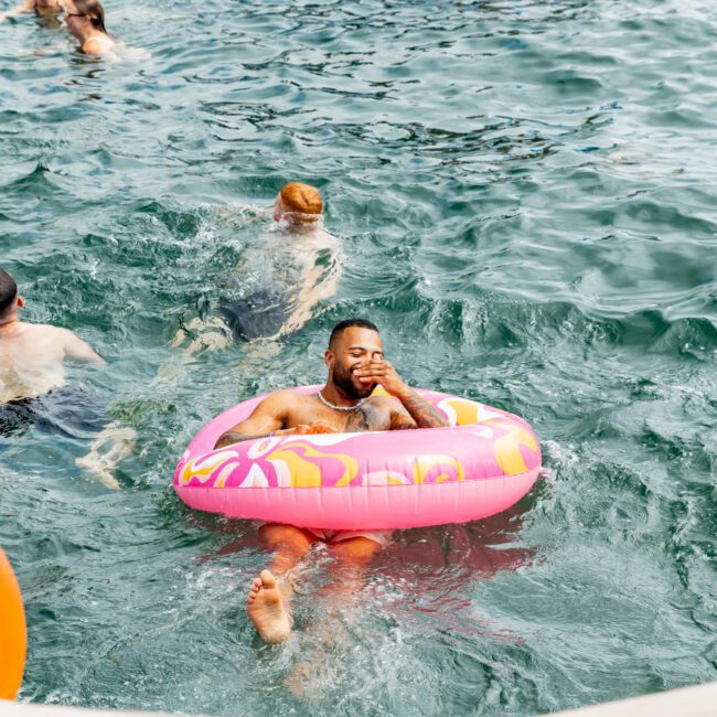 A man relaxes in a pink inflatable pool float on open water, surrounded by other people swimming and enjoying themselves under the bright sunlight.