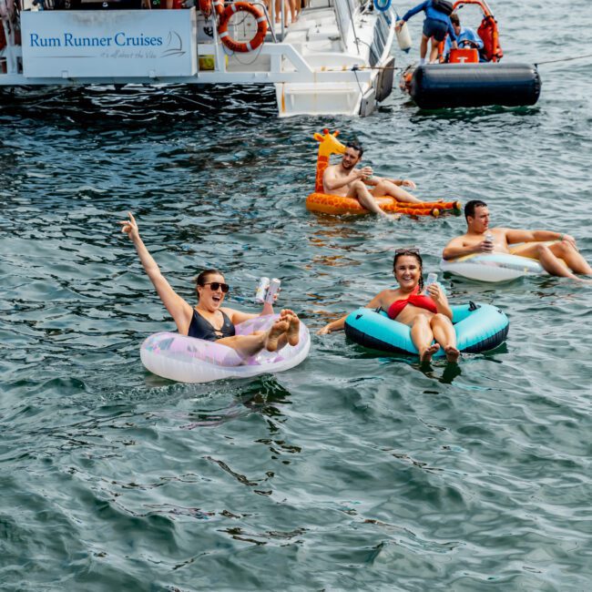 People relax on colorful floaties in the water near a boat labeled "Rum Runner Cruises." One woman points and holds a drink, while others smile. The city skyline is visible in the background on a sunny day.