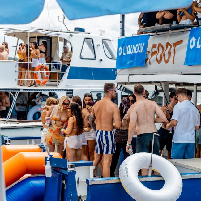 A group of people in swimsuits and summer clothing socializing and partying on docked boats, with bright umbrellas and an inflatable slide visible, under a partly cloudy sky.