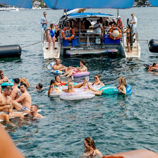 People relax and socialize on inflatable floats and swim in the water near a large anchored boat, with more people on the boat and city buildings visible in the background. The Yacht Social Club logo is at the bottom right.