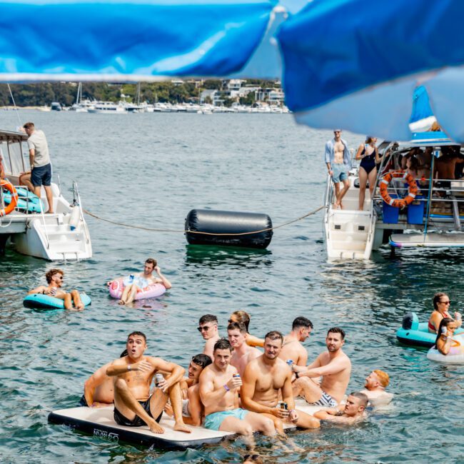 A group of young men sit closely together on a paddleboard in the water, surrounded by people on floaties and boats at a lively lakeside party. Blue umbrellas and partygoers are visible in the background.