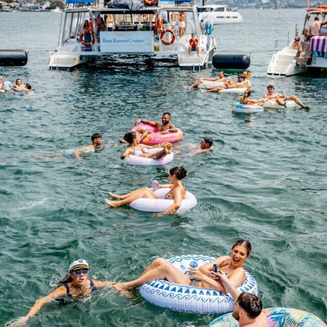 People float on colorful inflatable tubes in the water near a boat party. The boat is crowded with people dancing and enjoying music, with a city skyline visible in the background. The atmosphere is lively and festive.