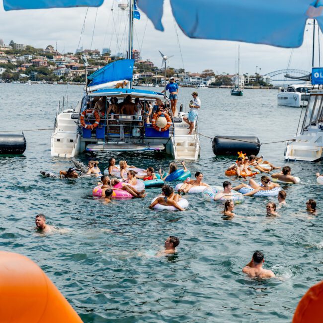 A group of people in colorful floaties and swimsuits enjoy a party in the water near docked boats on a sunny day. Blue umbrellas and an anchored yacht are in the foreground. Coastal buildings are visible in the background.