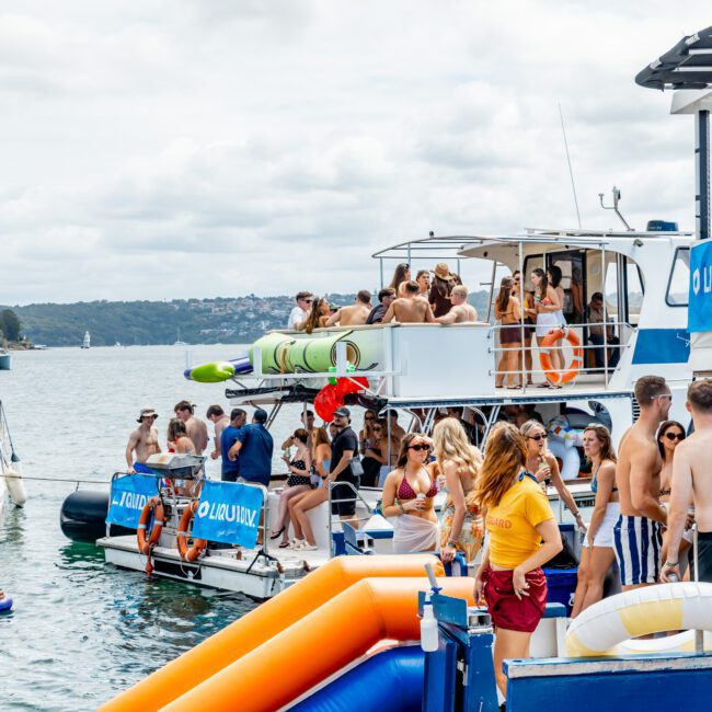 People enjoy a lively boat party on a sunny day, socializing on two connected boats with a blue and orange inflatable slide leading into the water. Many are in swimwear, and the atmosphere is festive and relaxed.