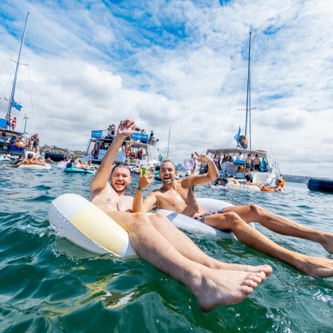 Two people float on an inflatable tube in clear water, smiling and waving at the camera. Behind them, several boats and groups of people enjoy a lively, sunny day on the water under a partly cloudy sky.