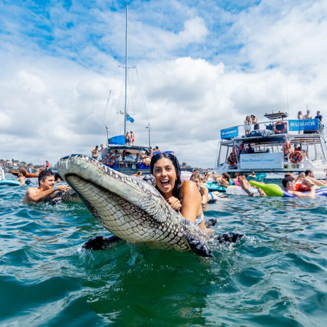 A woman in the water hugs a large inflatable crocodile while smiling, surrounded by people on floaties and boats during a lively water party under a partly cloudy sky.