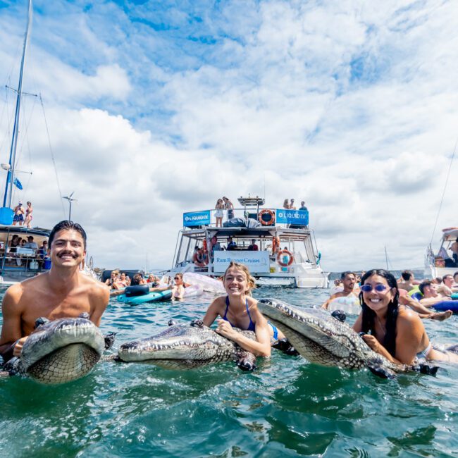 Three smiling people stand waist-deep in water holding inflatable crocodiles, with boats and groups of people relaxing on floaties in the background under a partly cloudy sky.
