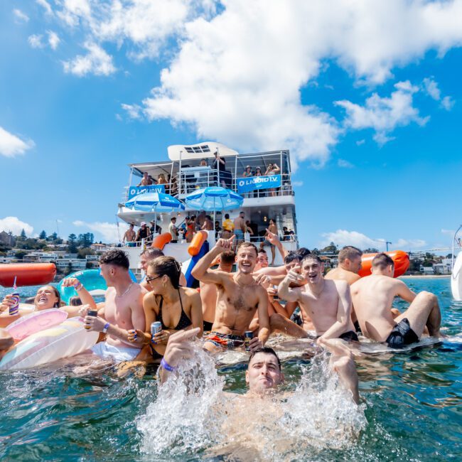 A group of young adults enjoy a lively pool party in the ocean, surrounded by large boats. Some float on inflatables, while others laugh and splash in the water under a sunny, blue sky.