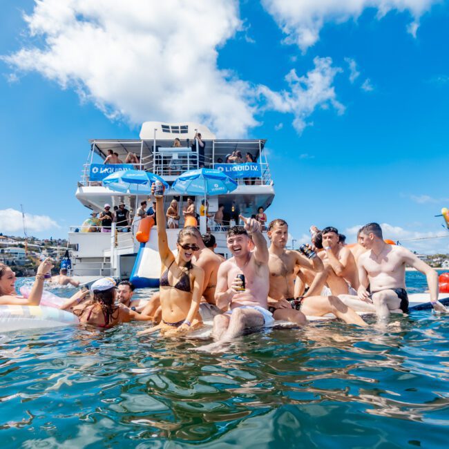 A group of people in swimsuits laugh and relax on inflatable floats in the water near a docked party boat, with bright blue skies and other boats in the background.